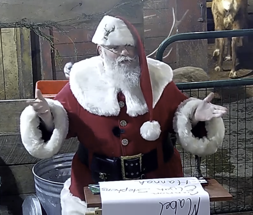 Santa holds arms out with Nice List scroll on table in front of him